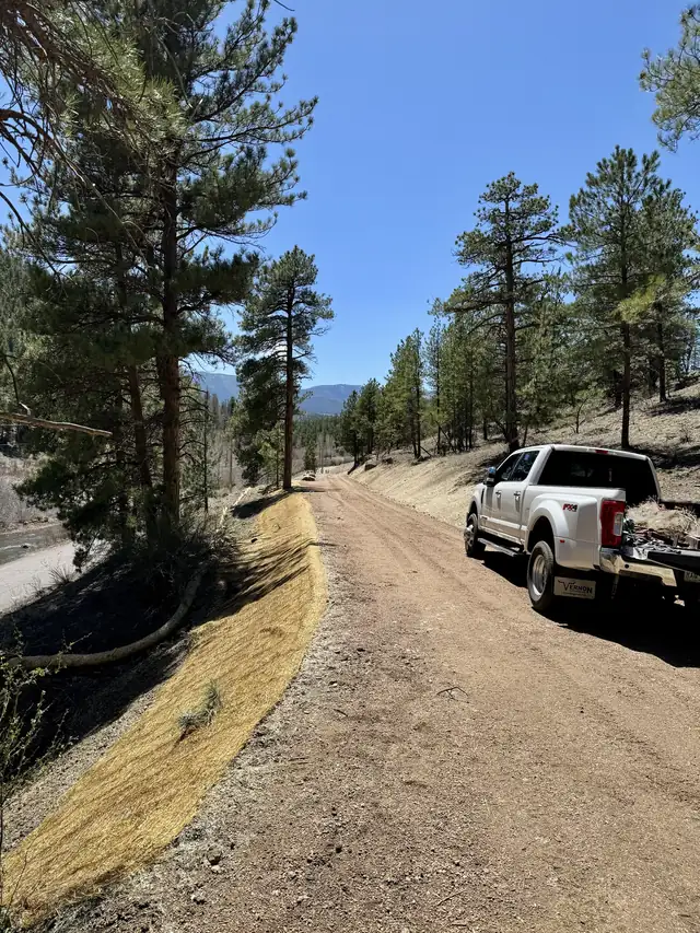 Finished graded mountain road through ponderosa pines with erosion control blanket along the slope edge beside a creek in Conifer, Colorado